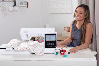 A woman sits at a table with a sewing and embroidery machine, fabric under the needle, and several spools of thread nearby.