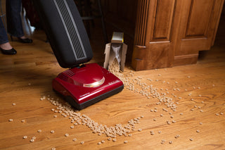 A red vacuum cleaner in use, picking up spilled cereal from a hardwood floor, next to an overturned cereal box.