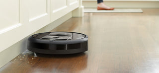 A robotic vacuum cleaner is cleaning debris near the base of a kitchen cabinet, with a person's foot visible in the background.