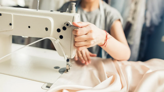 A person operates a sewing machine, guiding a piece of light-colored fabric under the needle in a workspace.