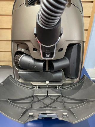 Close-up of a Sew & Vac C3 Miele Brilliant certified reconditioned vacuum cleaner base with attached hose and various settings visible, set against a blue striped background.