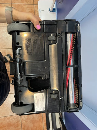 A person is holding the bottom of a Sew & Vac Riccar R10S vacuum cleaner, showcasing its metal brush roll and wheels against a tiled floor with a blue wall in the background.