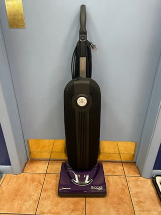A black Riccar R10S upright vacuum cleaner with a purple base stands against a blue wall on a tiled floor. The label reads "Sew & Vac," and it features a metal brush roll with 2 speeds for versatile cleaning.
