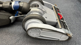 Close-up of the underside of the Oreck Magnesium RS Swivel-Steering Upright Vacuum RLW1500RS Certified Reconditioned, showing its wheels, brush, and belt on a carpeted surface.