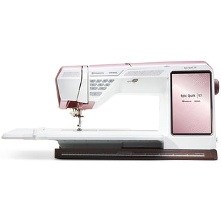 The Husqvarna Viking Epic Quilt 97 with Quilting Kit from Imagine Sewing & Vacuum is a computerized sewing and quilting machine featuring a large work area, digital display, control buttons, and white with pink accents.