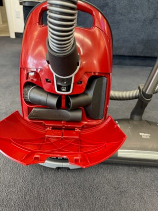 A Used Miele Complete C3 Canister Vacuum from Sew & Vac sits on a dark carpet with its front compartment open, showing stored attachments, hose, and premium power head—great for powerful cleaning and HEPA filtration.