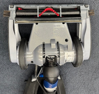 The Oreck Magnesium RS Swivel-Steering Upright Vacuum RLW1500RS features two large wheels, a roller brush with red bristles, and a gray plastic housing on the underside, shown here on a carpeted floor.