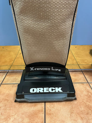 An Sew & Vac Oreck XL21 commercial upright vacuum cleaner on a carpeted floor with a blue wall in the background, featuring a new rubber belt.