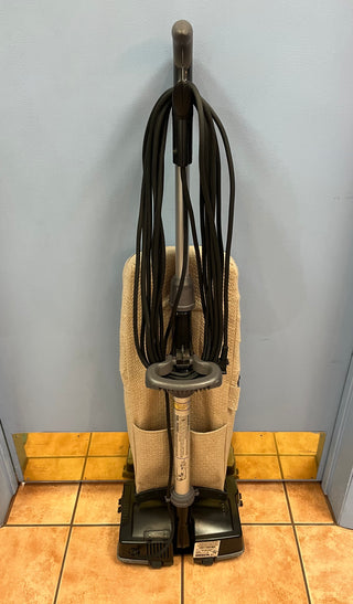 Sew & Vac's Oreck XL21 upright vacuum cleaner with attached hose and accessories, including a rubber belt, standing against a wall on a tiled floor.