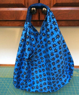 Blue Origami Market Bag crafted from fabric, with black handles featuring a pattern of star-like flowers, placed on a wooden surface with a green cutting mat below. (Sew & Vac)