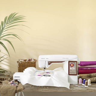 The Husqvarna Viking Designer Topaz 40 sewing and embroidery machine stitches on white fabric, surrounded by thread spools, rolls of fabric, and a potted plant on a wooden table.