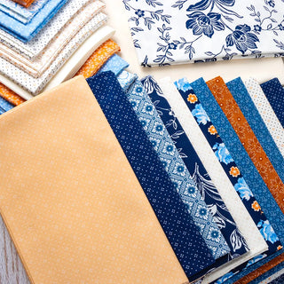 Stacks of blue, white, beige, and orange fabrics with floral, geometric, and polka dot prints from the Denim & Daisies collection are featured in the Denim Halloween Figs Quilt Kit by Box Kits.