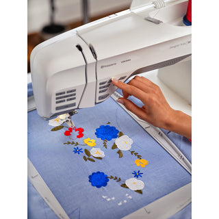 A person uses the Husqvarna Viking Designer Ruby 90 by 38-SVP - Singer Viking Pfaff to stitch colorful floral patterns with advanced embroidery features onto blue fabric.