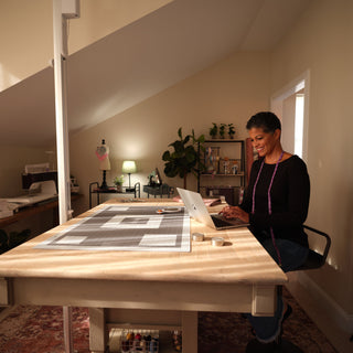 In a bright craft room, a person uses their laptop at a spacious worktable with fabric and tools, exploring customizable and paperless patterns for their next project with the Singer Ditto Pattern Projector.