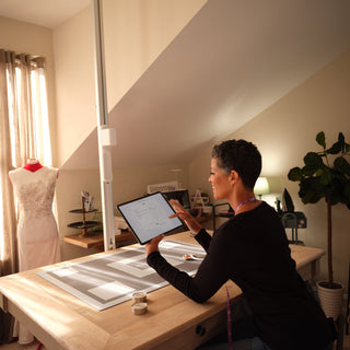 In a sewing room, a person uses a tablet at their worktable with a Singer Ditto Pattern Projector, surrounded by sewing supplies and a dress form with a beaded gown—showcasing customizable patterns for unique garment designs.