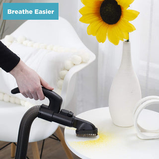 A hand uses the simplicity Simplicity S20EZM Allergy Bagged Upright vacuum’s brush attachment to clean yellow powder from a white table beside a chair and a vase with a sunflower.