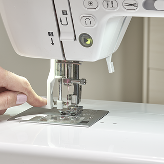 A close-up shows a person's hand guiding fabric under the needle of the Baby Lock Allegro Quilting and Sewing Machine by Baby Lock, preparing to sew.