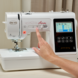 A person is using their right hand to press a button on the control panel of a Baby Lock Aurora Embroidery and Sewing Machine by Baby Lock. The machine, featuring 303 built-in embroidery designs, is positioned on a wooden surface with beige curtains in the background.