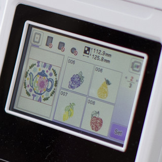 Close-up of a digital screen on the Baby Lock Bloom Sewing and Embroidery Machine by Baby Lock, showcasing five built-in designs such as a teapot, grapes, a pear, a lemon, and an apple.