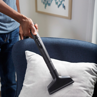 A person uses the simplicity Jill Canister Vacuum Cleaner to clean a white pillow on a blue upholstered chair.