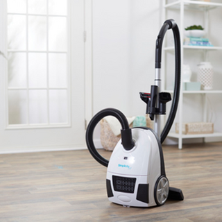 The simplicity Jill Canister Vacuum Cleaner with hose sits on a wooden floor in a bright, tidy room featuring a window and shelving in the background.