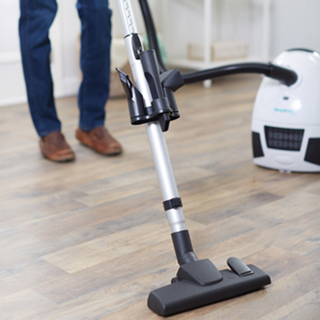 A person uses the Simplicity Jill Canister Vacuum Cleaner, featuring powerful suction, a metal wand, and a floor attachment to clean a wooden floor.