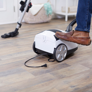 A person steps on a simplicity Jill Canister Vacuum Cleaner, a lightweight model with powerful suction and a disconnected power cord, on a wood floor.