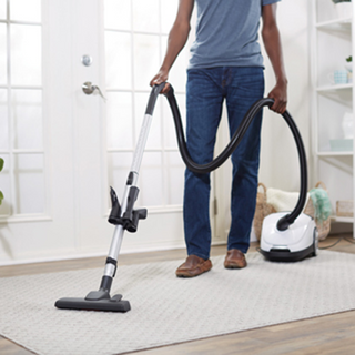 Wearing jeans and a t-shirt, a person uses the simplicity Jill Canister Vacuum Cleaner with strong suction to clean a light rug in a bright, tidy room.