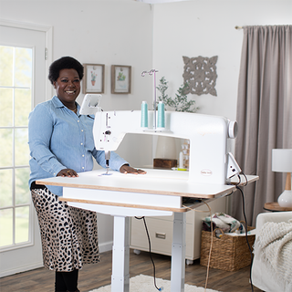 A smiling woman stands next to a Baby Lock 20" Regalia Stationary Machine With Table And Insert on a table in a bright, well-organized room.