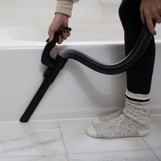 Wearing socks, a person uses the Simplicity S20EZM Allergy Bagged Upright vacuum by simplicity with a hose attachment to clean along the edge of a bathtub on a tiled floor.