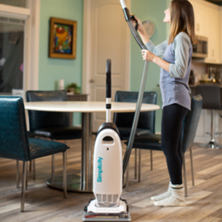A woman vacuums a hardwood floor in a dining area with a round table and four chairs using the simplicity Simplicity S20EZM Allergy Bagged Upright, which offers powerful suction and HEPA filtration.