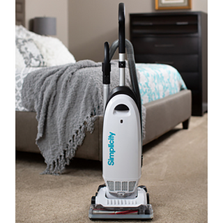 The Simplicity S20EZM Allergy Bagged Upright by simplicity stands on a beige carpet in a bedroom with a bed, gray blanket, and dark dresser in the background, delivering powerful suction for thorough cleaning.