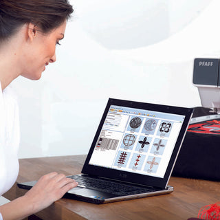 A woman works at a desk on embroidery designs using PFAFF Creative 3.0 and its Exclusive Stitch Creator software on her laptop, with an embroidery machine in the background.