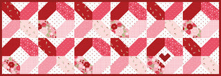 A rectangular quilt with a geometric pattern in shades of red, pink, and white, featuring hearts and floral prints, arranged in symmetrical rows.