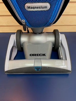 A reconditioned Oreck Magnesium LW1500RS vacuum cleaner, lightweight and sleek in design, sits atop a table with its vibrant blue and silver color scheme catching the eye.
