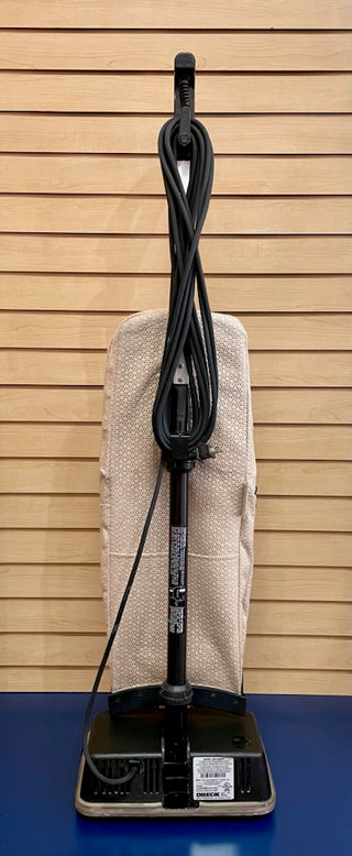 A Sew & Vac Oreck XL - Certified Reconditioned vacuum cleaner with a beige dust bag secured neatly around the handle, standing upright against a blue slatted wall.