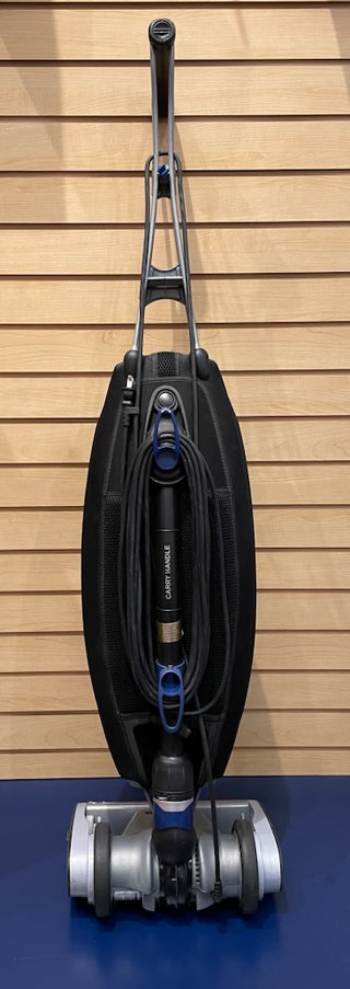 Sew & Vac's Oreck Magnesium - Certified Refurbished upright vacuum cleaner with a black oval-shaped dust compartment, mounted on a blue carpet in front of a wooden slat wall. This lightweight vacuum is in great condition.