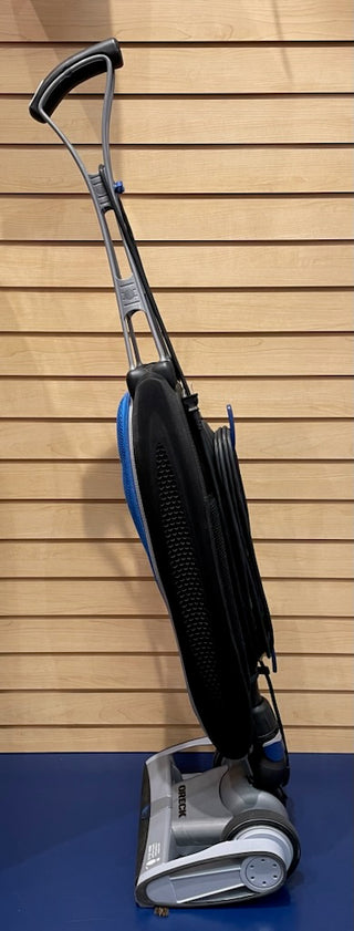 Sew & Vac's Oreck Magnesium - Certified Refurbished upright vacuum cleaner with a black and silver body, standing against a wooden slat wall.