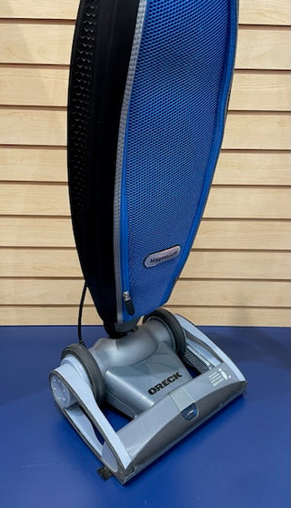 A reconditioned upright Sew & Vac Oreck Magnesium LW1500RS vacuum cleaner against a wall with a blue bag.