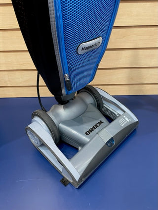 A certified refurbished Sew & Vac Oreck Magnesium LW1500 upright vacuum cleaner on a blue surface, showcasing its blue and gray body and compact design.
