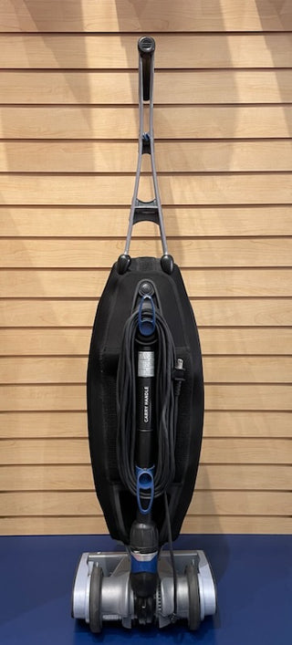 A Sew & Vac certified refurbished Oreck Magnesium LW1500 black upright vacuum cleaner against a wooden slat wall.