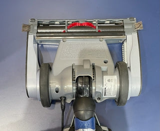Overhead view of a Oreck Magnesium LW1500 vacuum cleaner with visible brushes and a plugged-in power cord, set against a blue background.