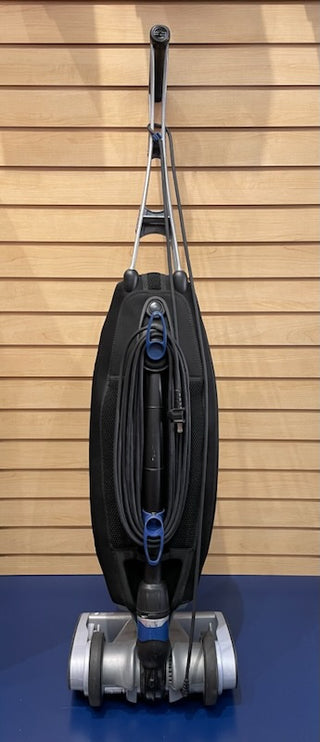Sew & Vac's Oreck Magnesium LW1500 - Certified Refurbished vacuum cleaner with a gray and black design, featuring an attached hose and mounted on a blue carpet against a wooden slat wall.