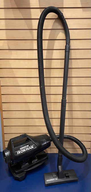 Upright Sew & Vac Oreck Ironman vacuum cleaner with strong suction and a flexible hose attachment, labeled "Ironman," standing against a wooden slat wall.