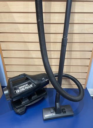 Sew & Vac Ironman vacuum cleaner with strong suction, featuring a flexible hose and floor attachment, standing upright against a wooden slat wall.