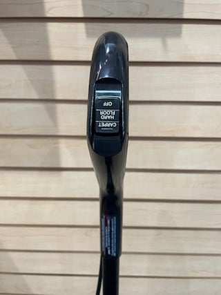 Close-up of a black bicycle handlebar with a Sew & Vac Oreck Elevate Cordless digital display showing battery status, against a hardwood background.