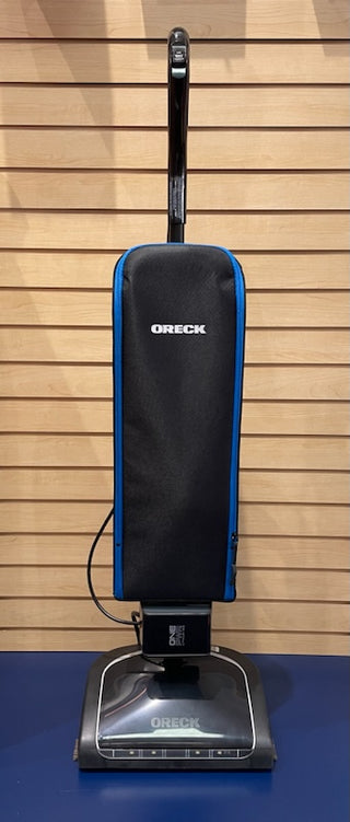 A reconditioned Sew & Vac Oreck Elevate Cordless upright vacuum cleaner with a black body and blue accents, standing against a wooden slat wall.
