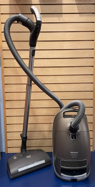A reconditioned Sew & Vac C3 Miele Brilliant vacuum cleaner with a silver canister and attached hose, positioned upright next to its floorhead against a wooden slat wall.