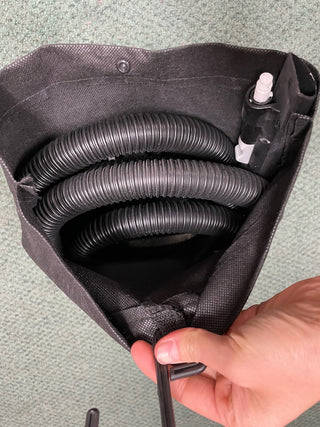 A person's hand holds open a Sew & Vac Certified Refurbished black bag with coiled cable tubing and a spray bottle inside containing the Hoover Smartwash Pet Complete Carpet Cleaner.