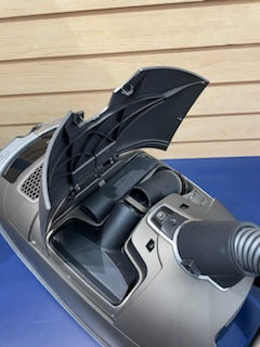 A certified refurbished Miele C3 Brilliant silver and black vacuum cleaner with an open dirt compartment, displaying its internal filter and structure, against a blue and wooden background.
